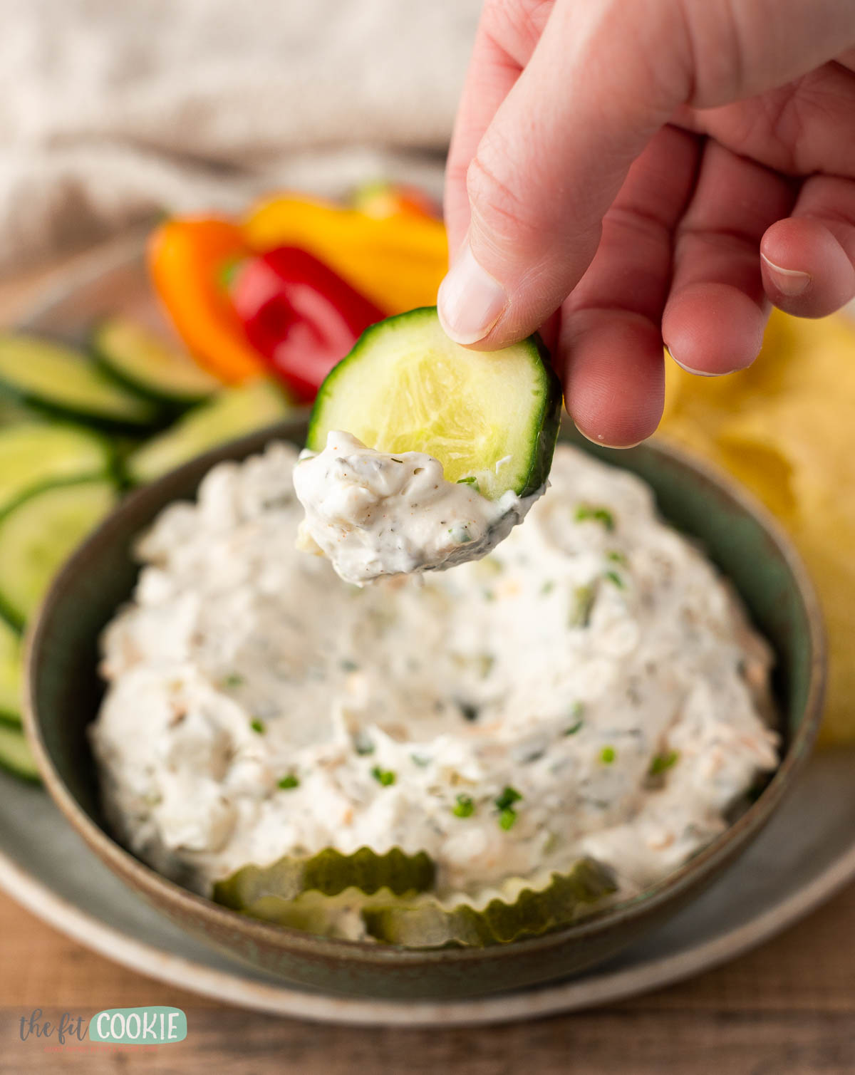 A hand holds a cucumber slice with dill pickle dip on top, with sliced vegetables and chips arranged next to the bowl.