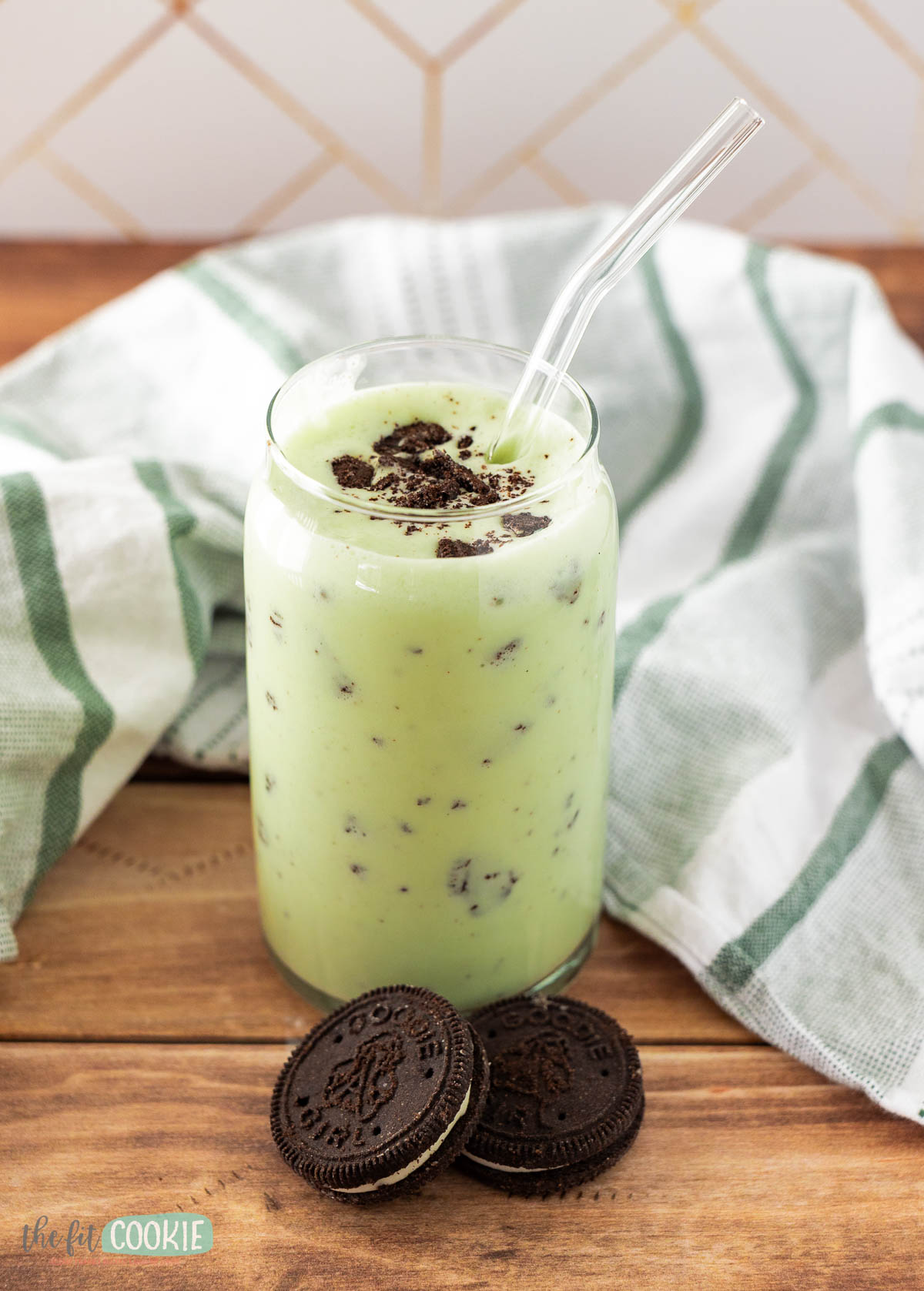 A glass of light green, dairy free shamrock shake with crushed cookies on top and a clear straw, placed on a wooden surface next to two chocolate sandwich cookies and a striped towel.