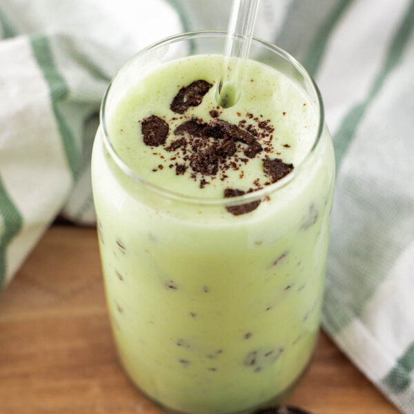 A glass of dairy free shamrock shake with a clear straw, topped with crushed chocolate cookies, sits on a wooden surface next to two whole cookies.