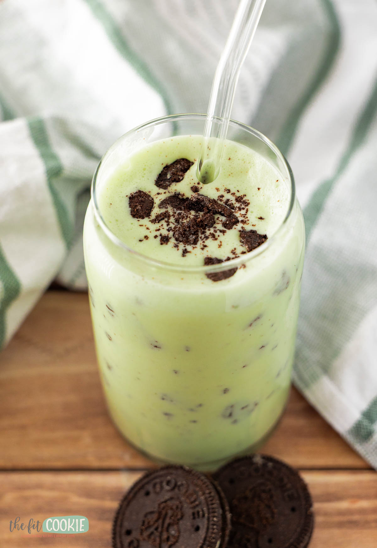A glass of dairy free shamrock shake with a clear straw, topped with crushed chocolate cookies, sits on a wooden surface next to two whole cookies.