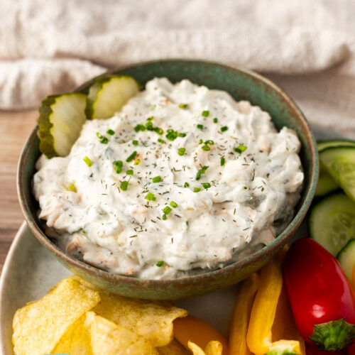 A bowl of creamy dill pickle dip garnished with chopped chives and dill, served with potato chips, pickles, cucumber slices, and sliced bell peppers.