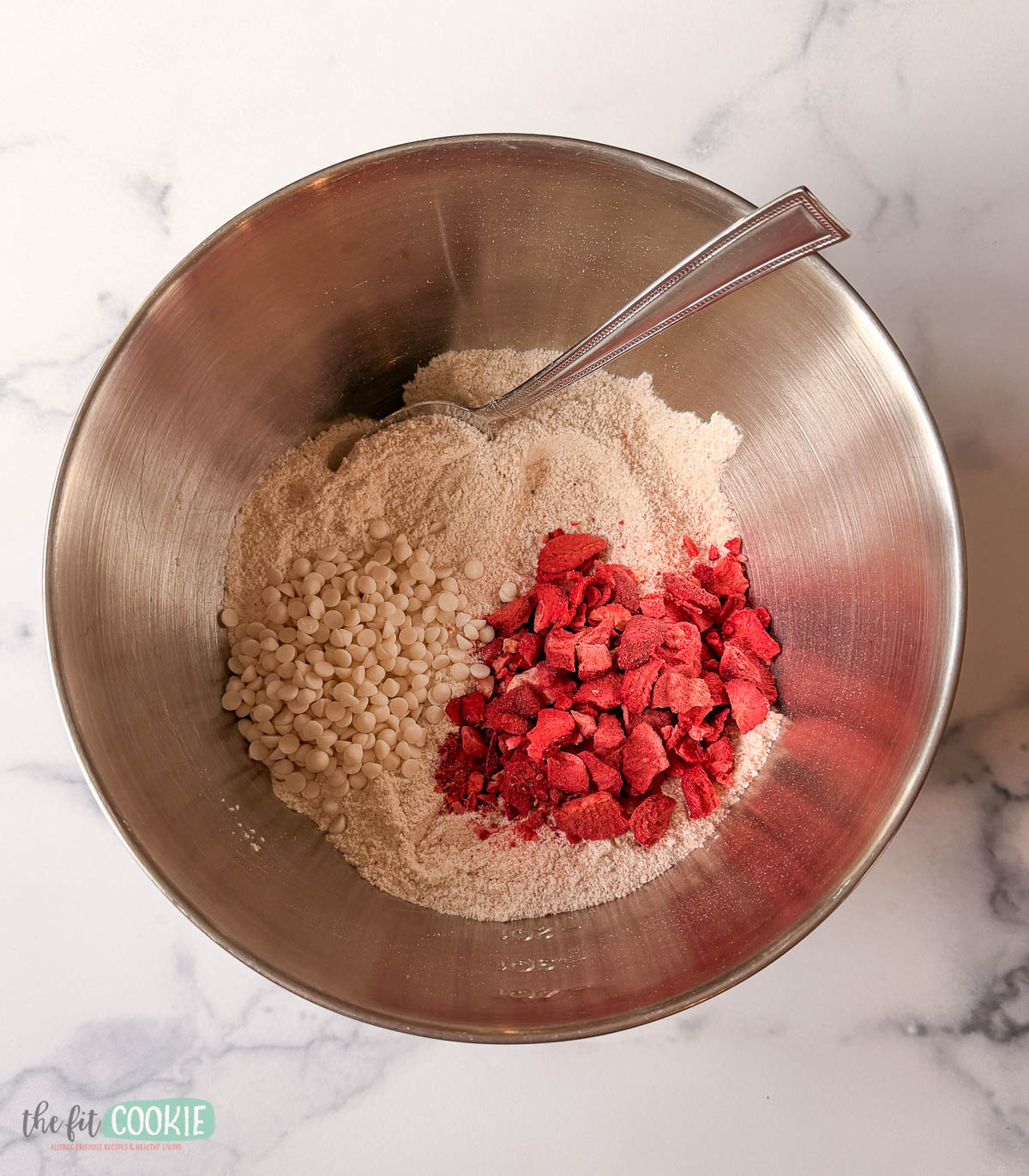 A metal mixing bowl with dry ingredients for strawberry blondies, including white chocolate chips, freeze-dried strawberries, and flour, with a metal spoon inside.