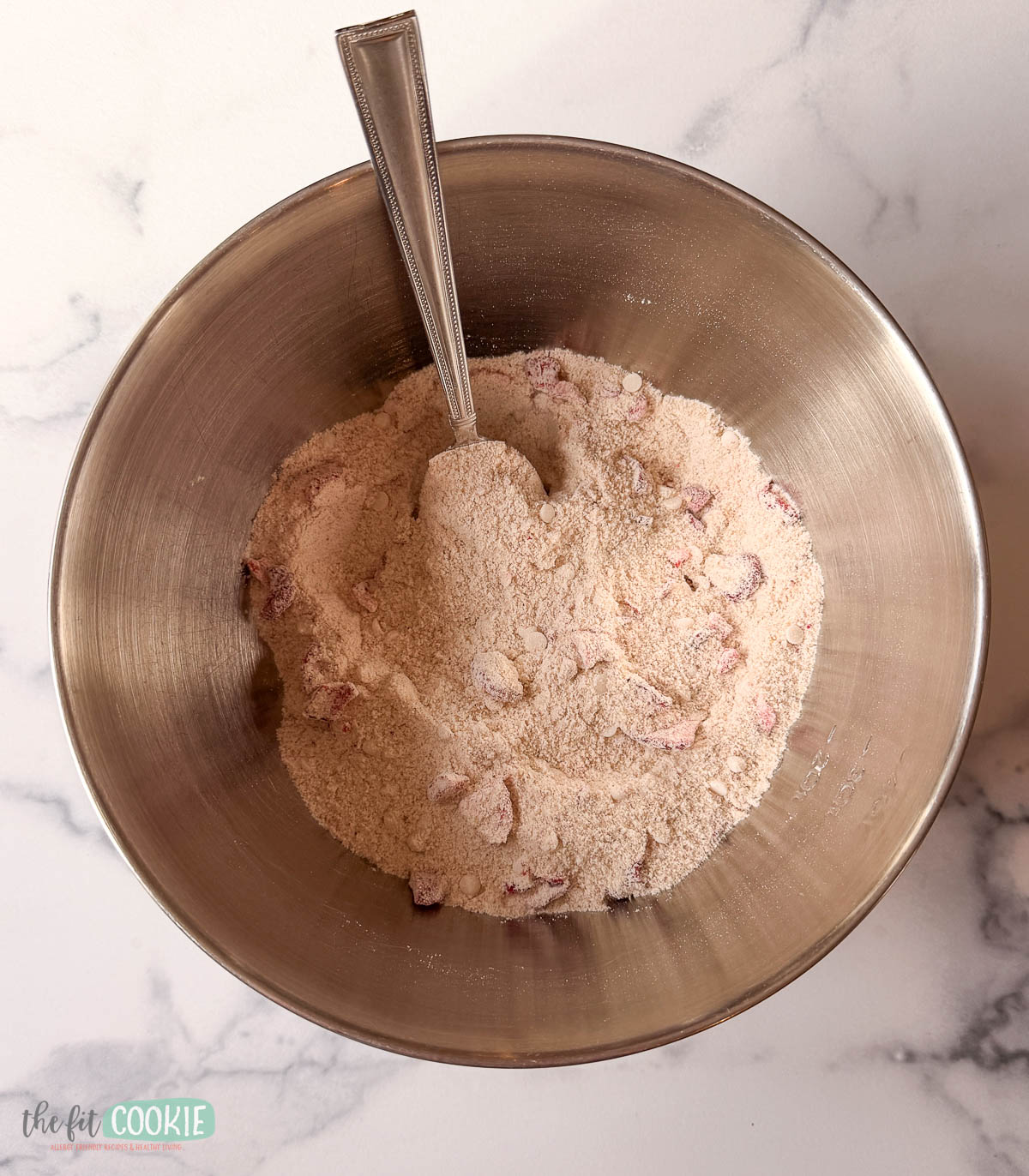 A metal mixing bowl with a spoon, containing dry baking mix for strawberry blondies with visible pieces of dried fruit, on a marble countertop.