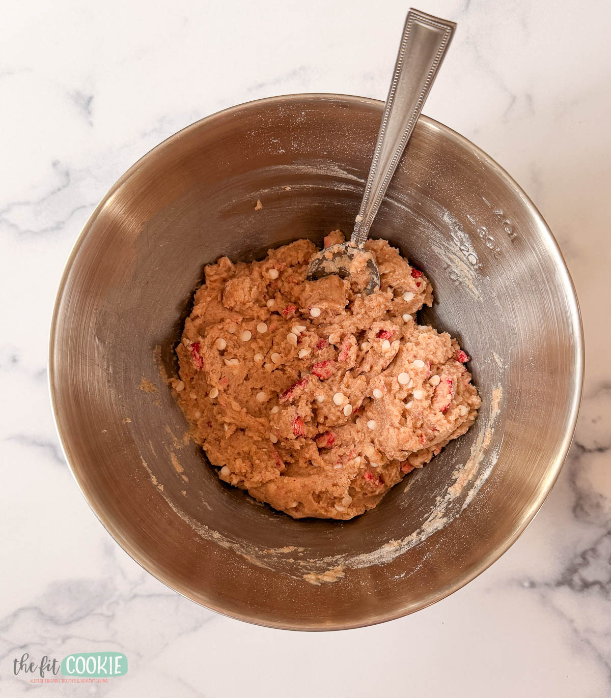 A metal mixing bowl with strawberry blondies cookie dough, filled with white and red mix-ins, and a metal spoon resting in the dough, on a marble countertop.
