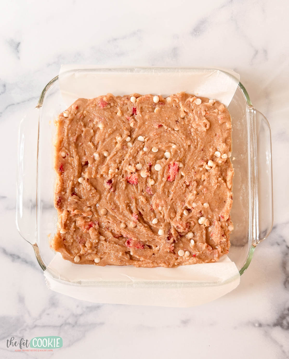 Square glass baking dish with unbaked strawberry blondies batter, mixed with white chocolate chips and red bits, lined with parchment paper on a marble surface.