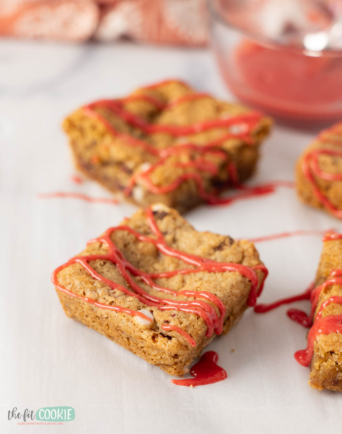Three square strawberry blondies topped with a red drizzle are arranged on a white surface, with a bowl of red sauce in the background.