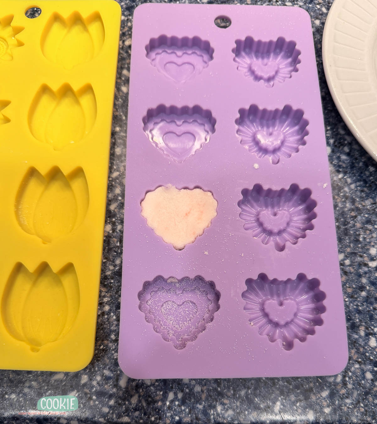 Purple silicone mold with heart and flower shapes, one filled with Dairy Free Cream Cheese Mints mixture, next to a yellow mold with tulip shapes.