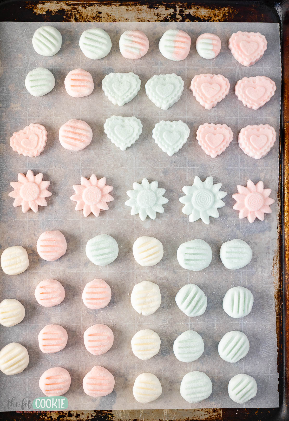 Rows of pastel-colored Dairy Free Cream Cheese Mints in various shapes—circles, flowers, and hearts—are arranged on a parchment-lined baking sheet.