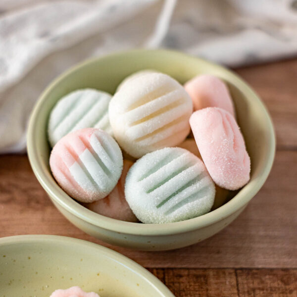 A small bowl filled with pastel-colored, round Dairy Free Cream Cheese Mints pressed with a striped pattern sits on a wooden surface.