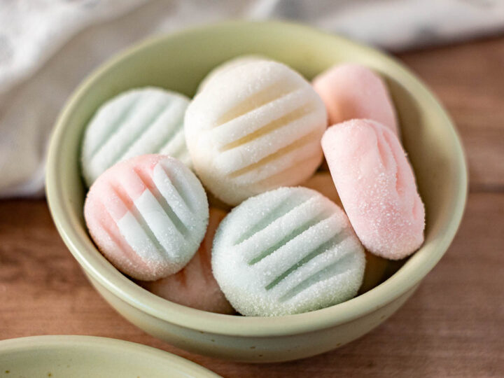 A small bowl filled with pastel-colored, round Dairy Free Cream Cheese Mints pressed with a striped pattern sits on a wooden surface.