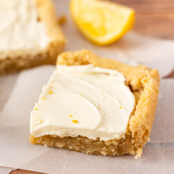 A square frosted lemon bar with creamy white frosting sits on parchment paper, with a lemon wedge in the background.