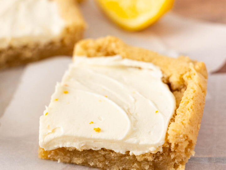 A square frosted lemon bar with creamy white frosting sits on parchment paper, with a lemon wedge in the background.