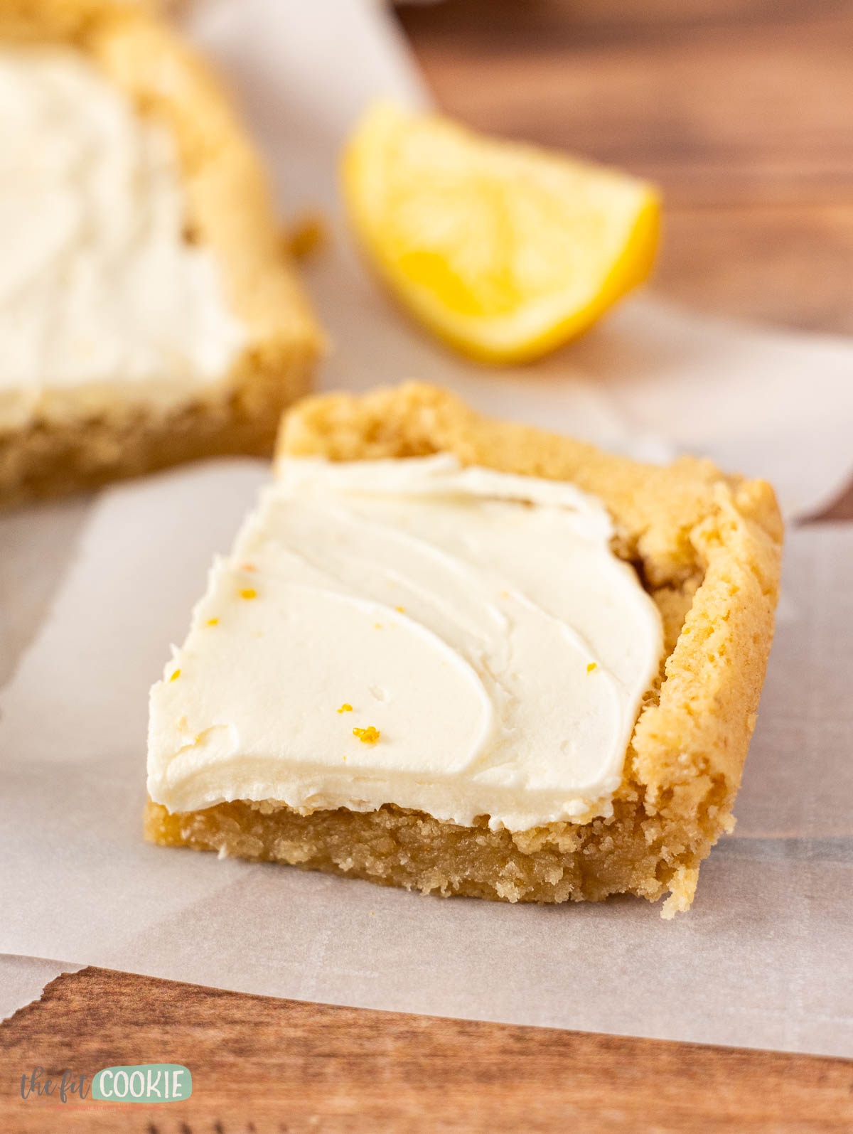 A square frosted lemon bar with creamy white frosting sits on parchment paper, with a lemon wedge in the background.