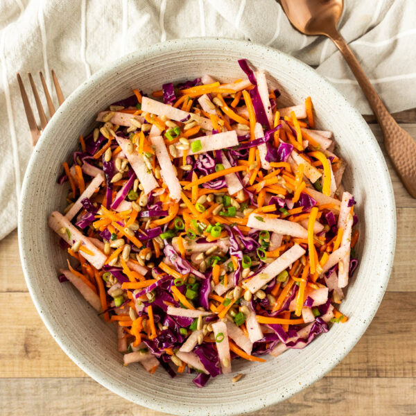 A bowl of vibrant jicama salad with shredded red cabbage, carrots, green onions, and sunflower seeds sits on a wooden table beside a fork, spoon, and a white cloth napkin.