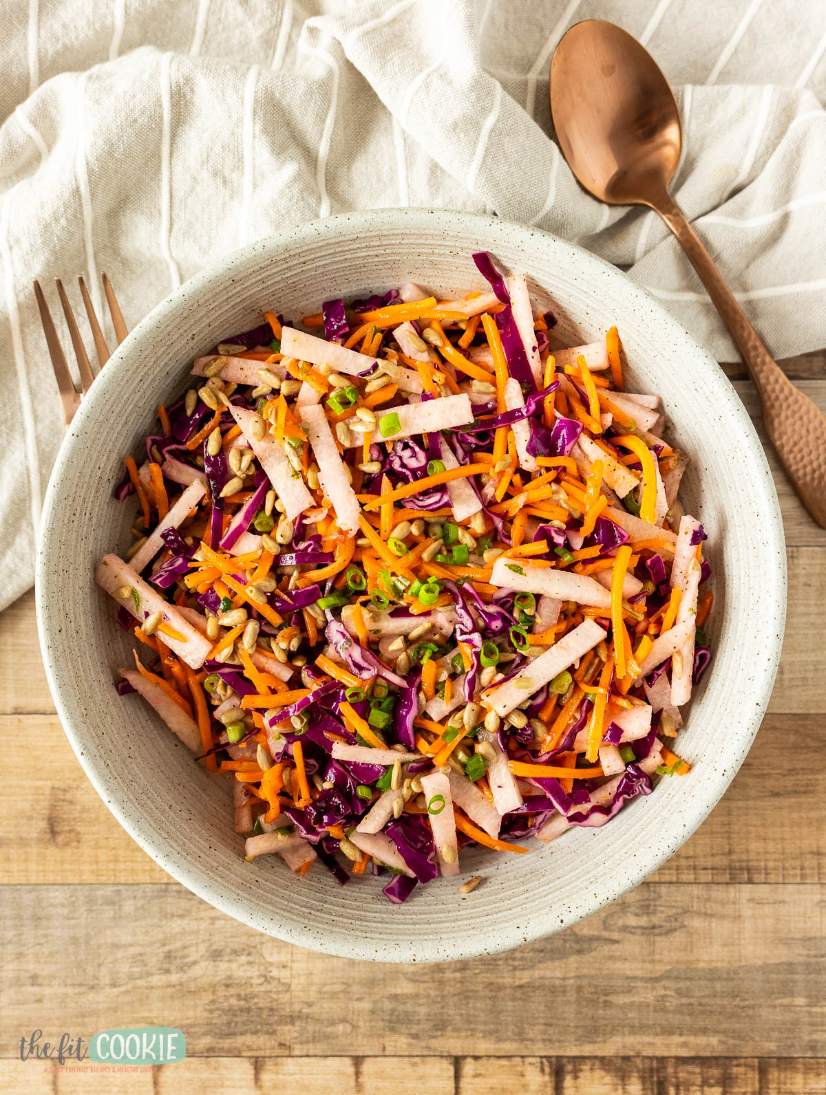 A bowl of vibrant jicama salad with shredded red cabbage, carrots, green onions, and sunflower seeds sits on a wooden table beside a fork, spoon, and a white cloth napkin.