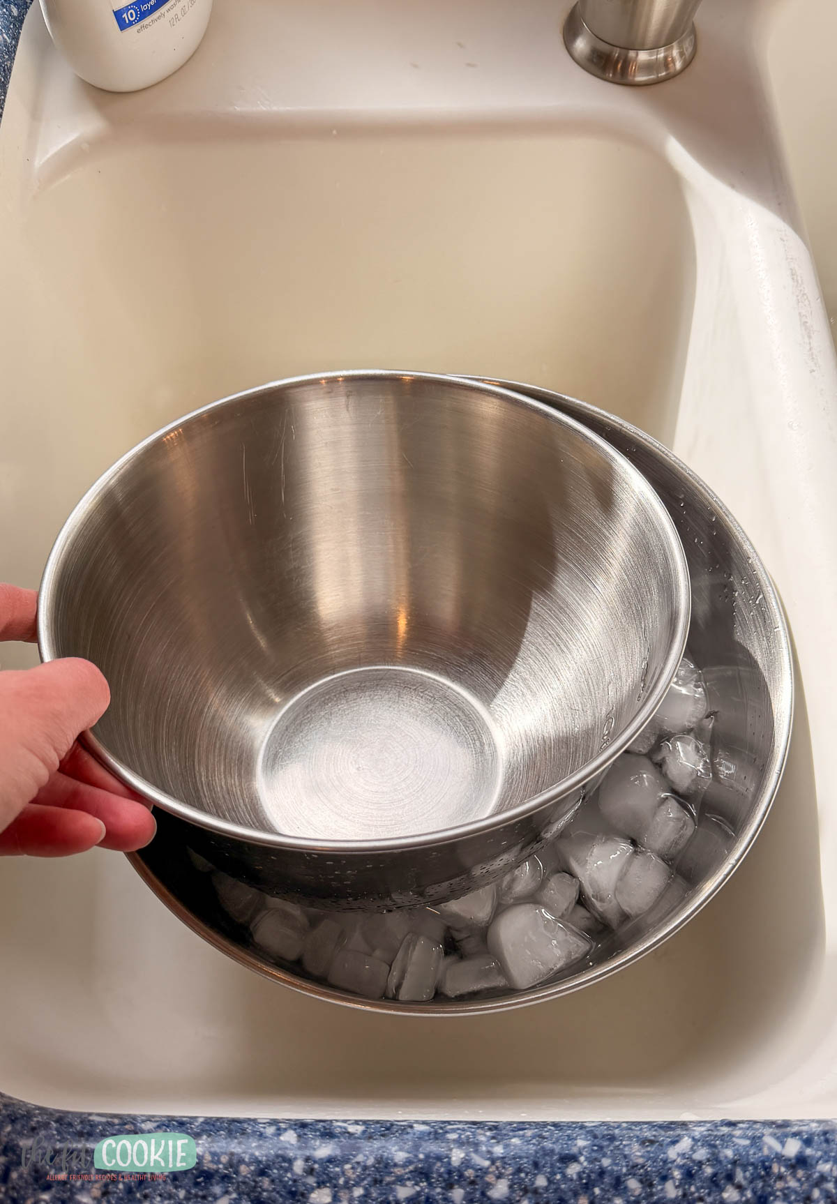 A hand places a metal mixing bowl into a larger bowl filled with ice cubes and water in a kitchen sink.