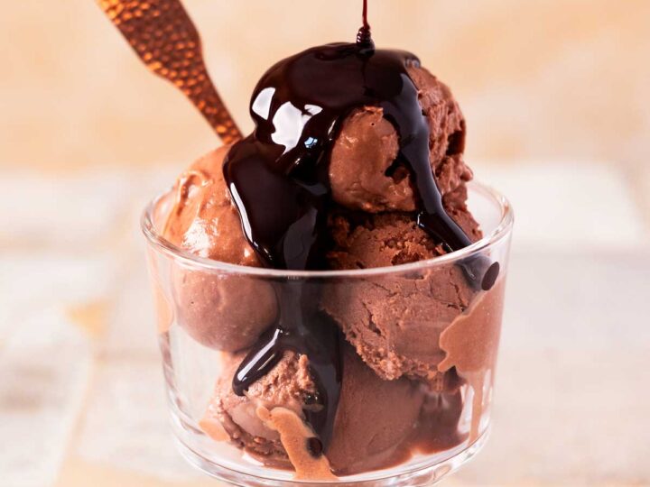 A glass dish filled with scoops of dairy free chocolate ice cream, topped with rich chocolate syrup, with a spoon placed inside.