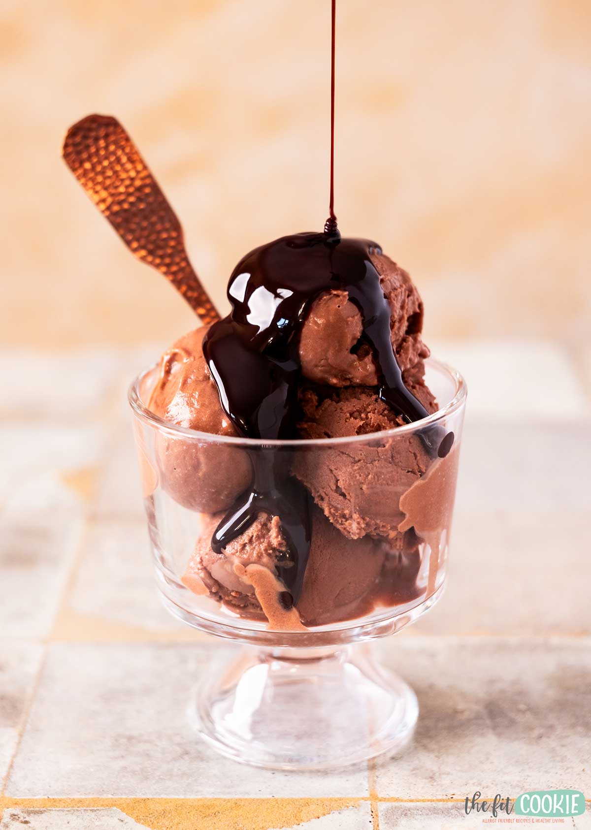 A glass dish filled with scoops of dairy free chocolate ice cream, topped with chocolate syrup, with a copper spoon placed inside.