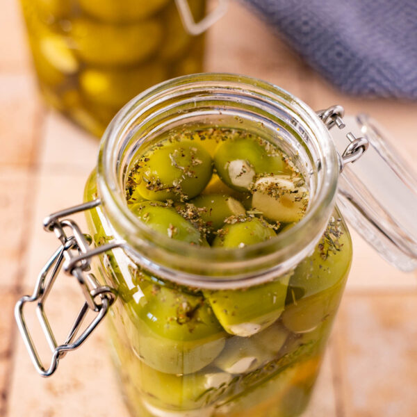 Glass jar filled with marinated olives, herbs, and liquid brine on a tiled surface. Another jar and a blue cloth are visible in the background. This snack is dairy free and packed with flavor.