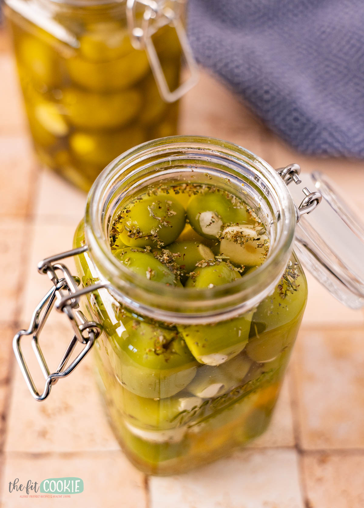 Glass jar filled with marinated olives, herbs, and liquid brine on a tiled surface. Another jar and a blue cloth are visible in the background. This snack is dairy free and packed with flavor.