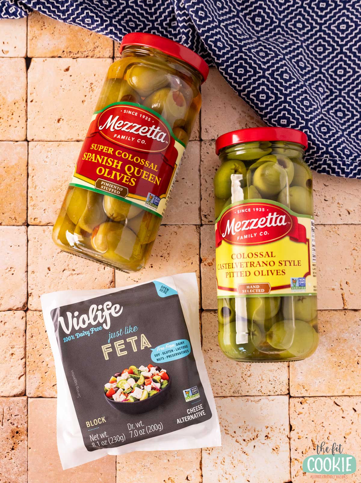 Two jars of Mezzetta olives and a package of Violife dairy free Just Like Feta cheese alternate are placed on a tiled surface.