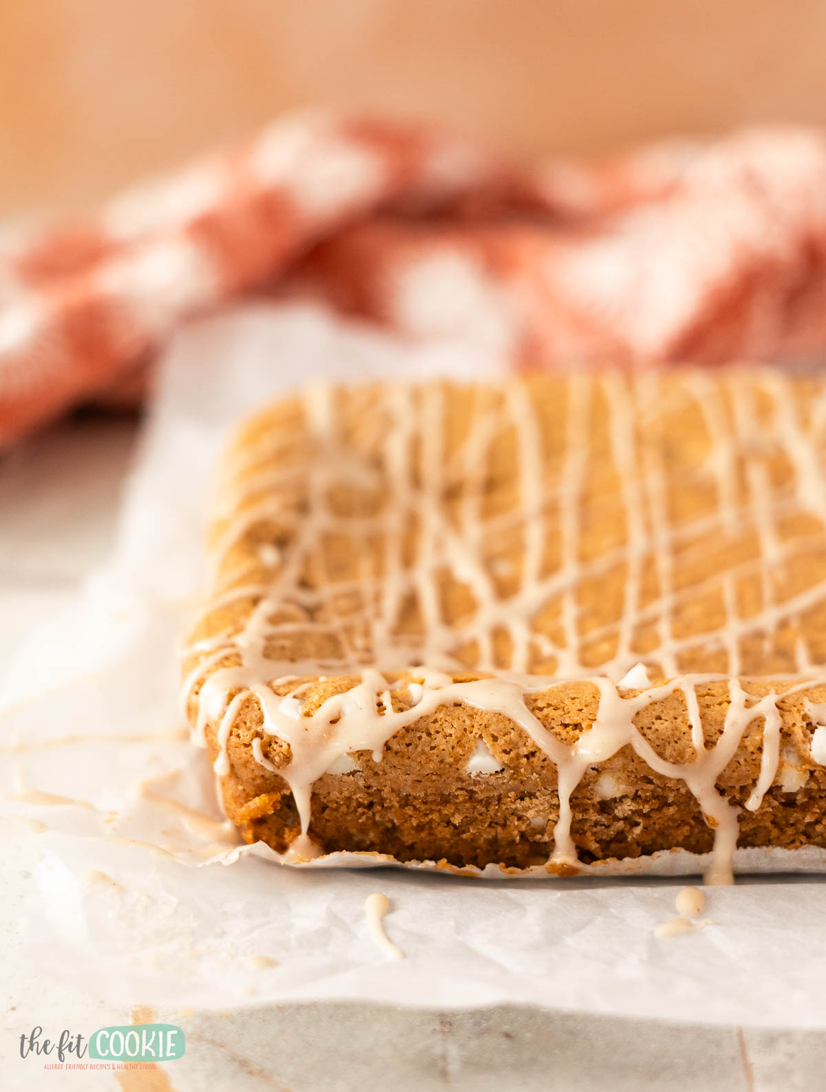 An uncut batch of vegan cinnamon roll blondies with white icing drizzle sits on parchment paper, with a red and white cloth in the background.