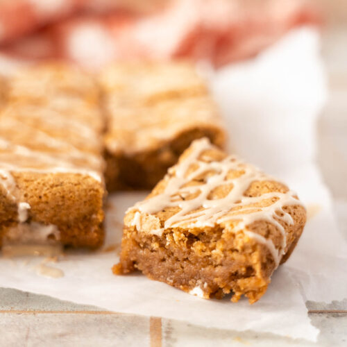 Close-up of glazed pumpkin bars on parchment paper, with one bar pulled slightly forward showing a moist interior—perfect for fans of cinnamon roll blondies.