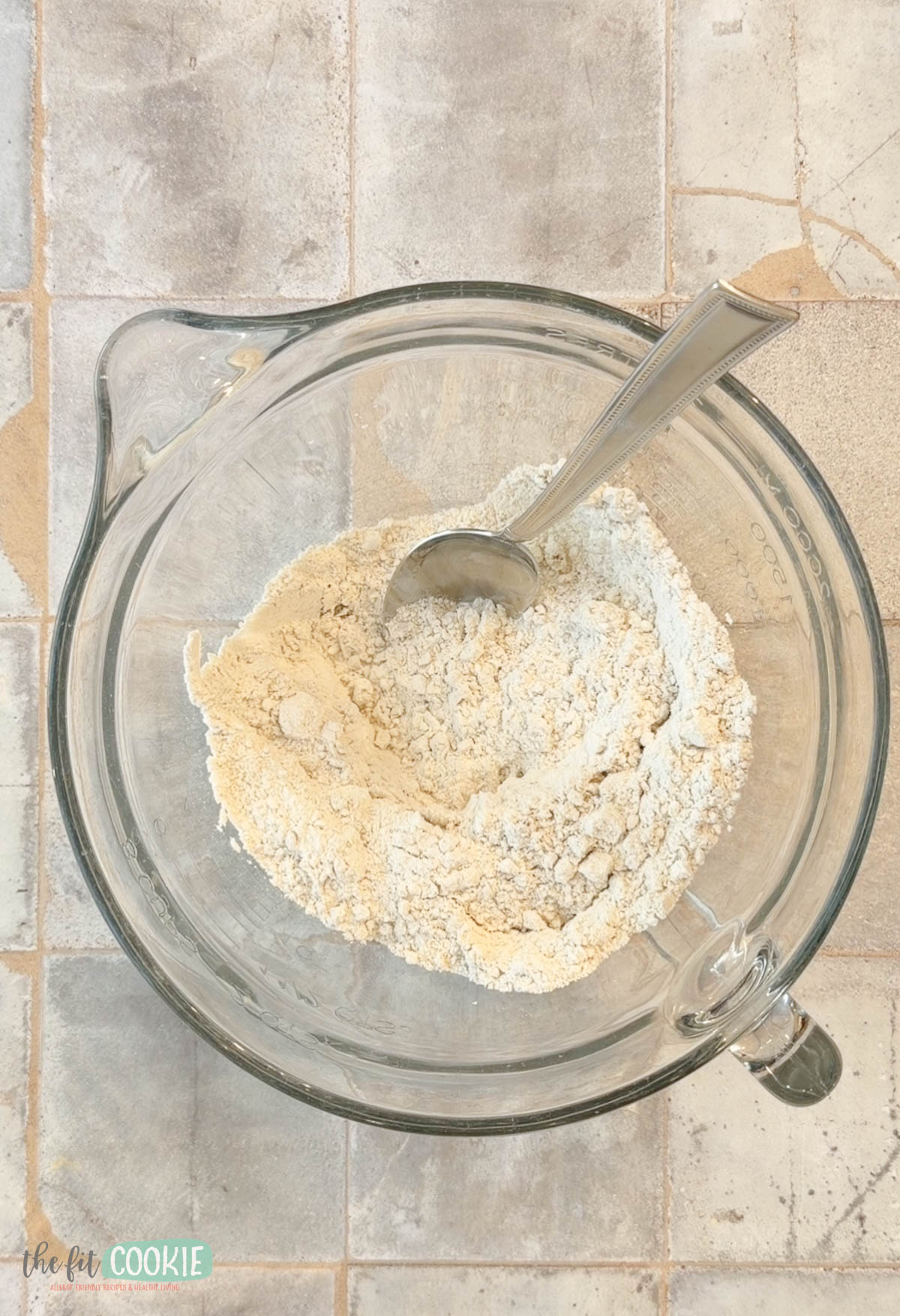 A glass mixing bowl with gluten free flour and a metal spoon inside.