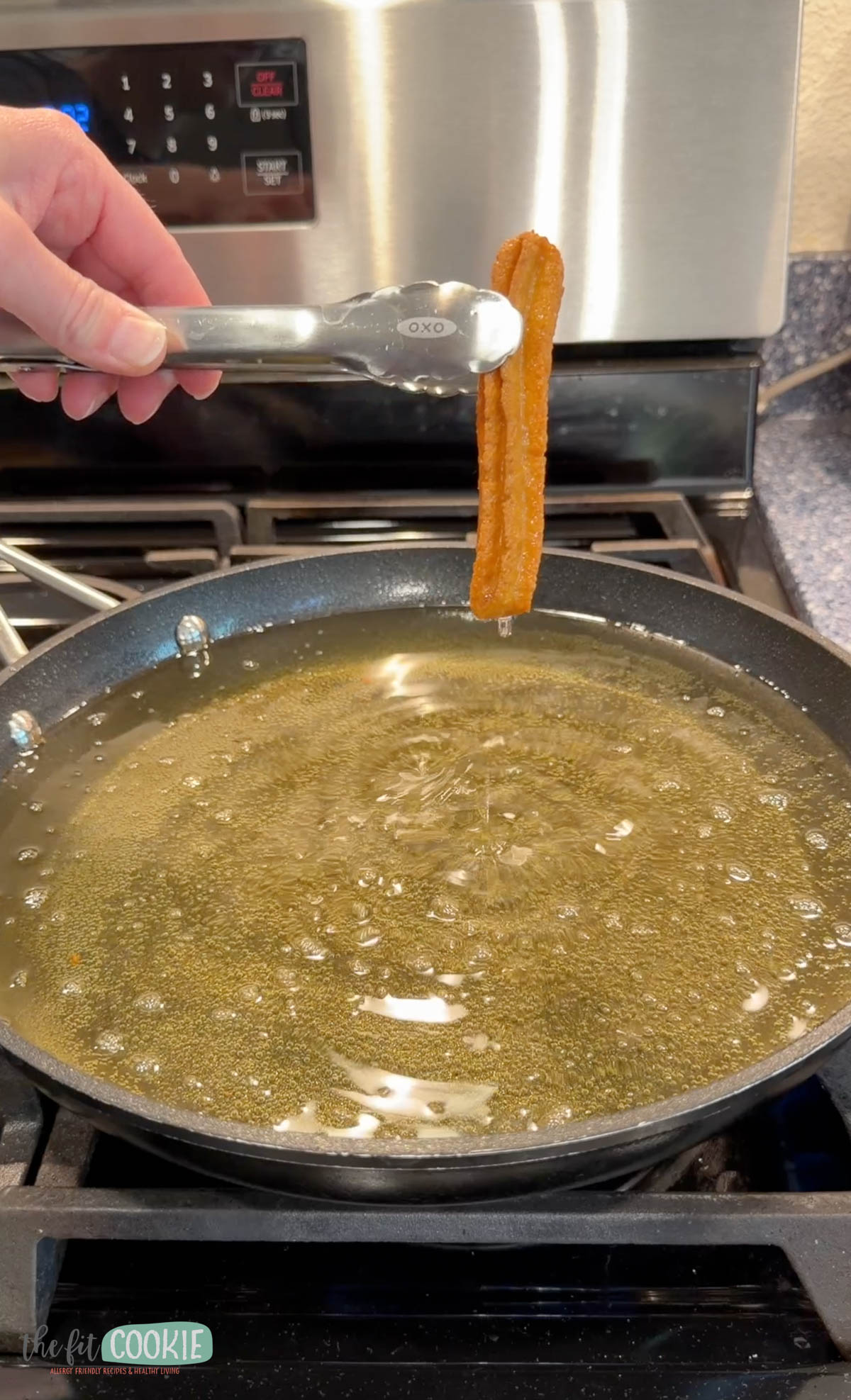 A hand holding metal tongs takes a vegan churro from a frying pan filled with hot oil on a stovetop.