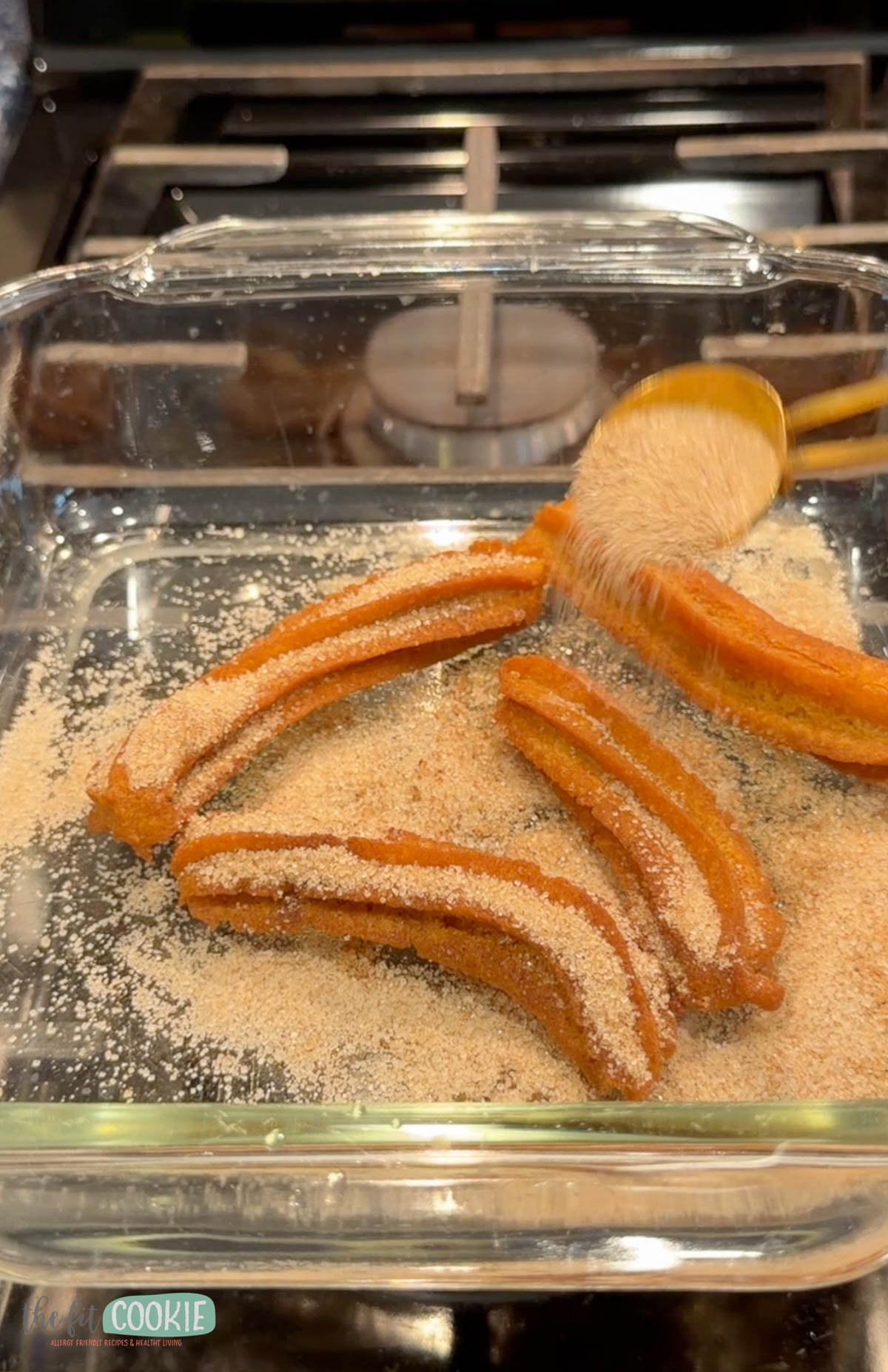 Four gluten free churros in a glass baking dish are being sprinkled with cinnamon sugar from a spoon.