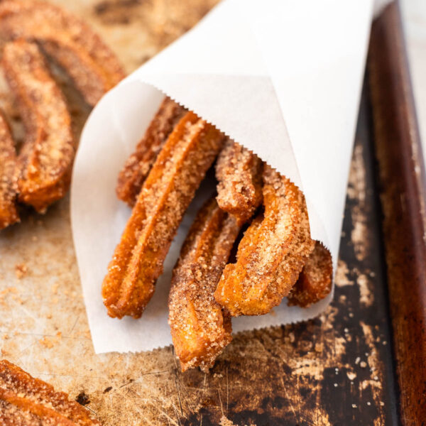 Several cinnamon sugar-coated Gluten Free Vegan Churros are wrapped in a white paper cone and rest on a baking sheet.
