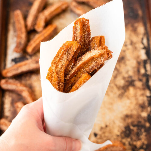 A hand holds a white paper cone filled with vegan churros coated in cinnamon sugar, with more churros on a baking sheet in the background.