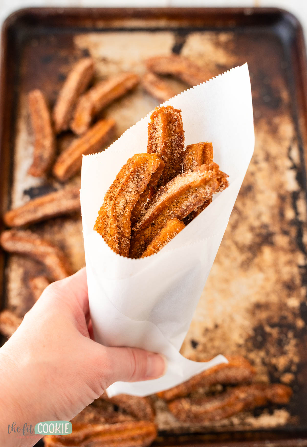 A hand holds a white paper cone filled with gluten free and vegan churros coated in cinnamon sugar.