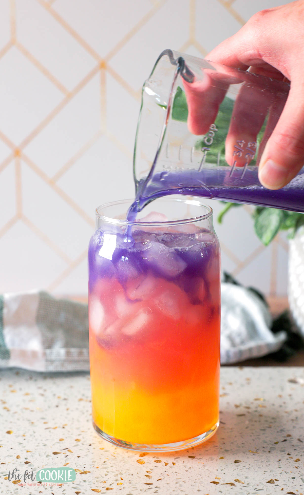 A hand pours purple liquid from a glass container into a clear glass filled with ice and yellow-orange liquid, creating a layered tropical butterfly drink.