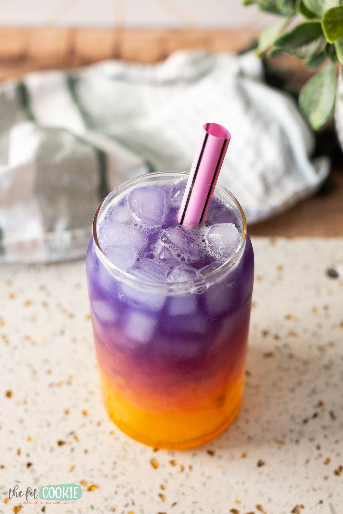 A glass filled with ice and a layered purple, pink, and orange Tropical Butterfly Drink, topped with a pink straw.