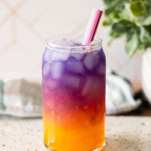 A glass filled with ice and a layered purple and orange Tropical Butterfly Drink, topped with a pink straw, sits on a speckled countertop with a leafy plant in the background—perfect for your next Starbucks Copycat homemade treat.