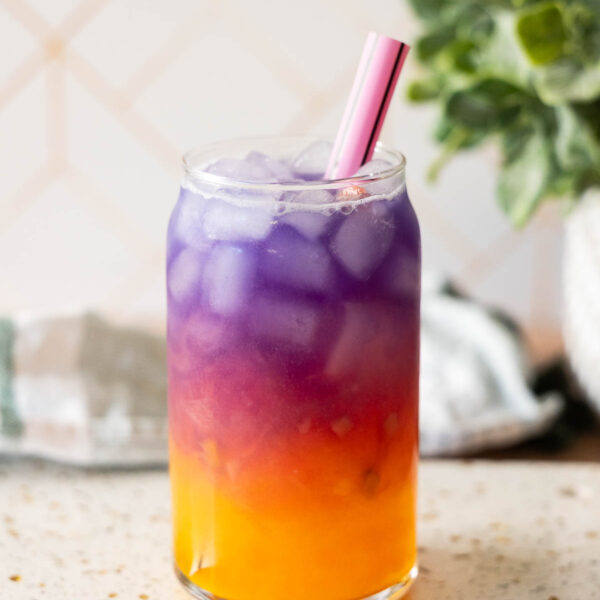 A clear glass filled with ice and a layered purple and orange Tropical Butterfly Drink, with a pink straw, sits on a speckled countertop near a green plant—a perfect homemade Starbucks copycat treat.