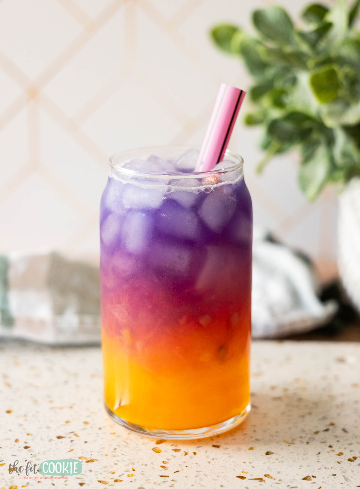 A clear glass filled with ice and a layered purple and orange Tropical Butterfly Drink, with a pink straw, sits on a speckled countertop near a green plant.