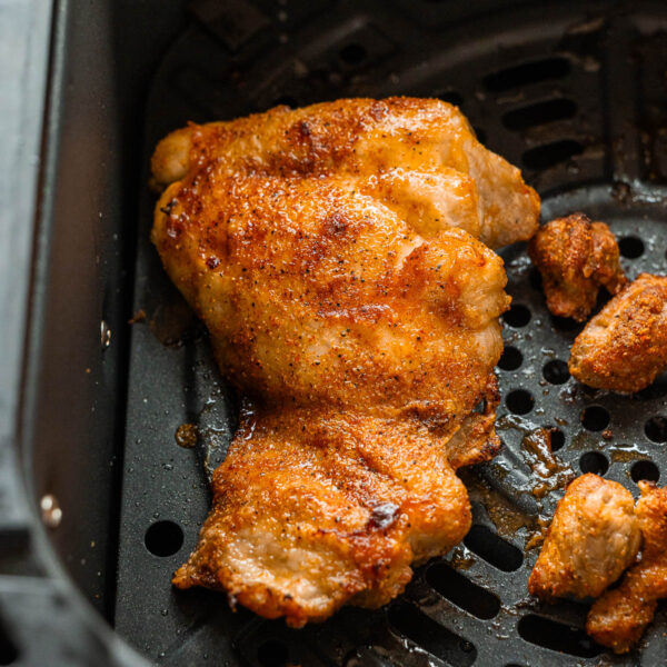 A cooked, seasoned air fryer chicken thigh sits in the basket with a few small pieces of chicken nearby.