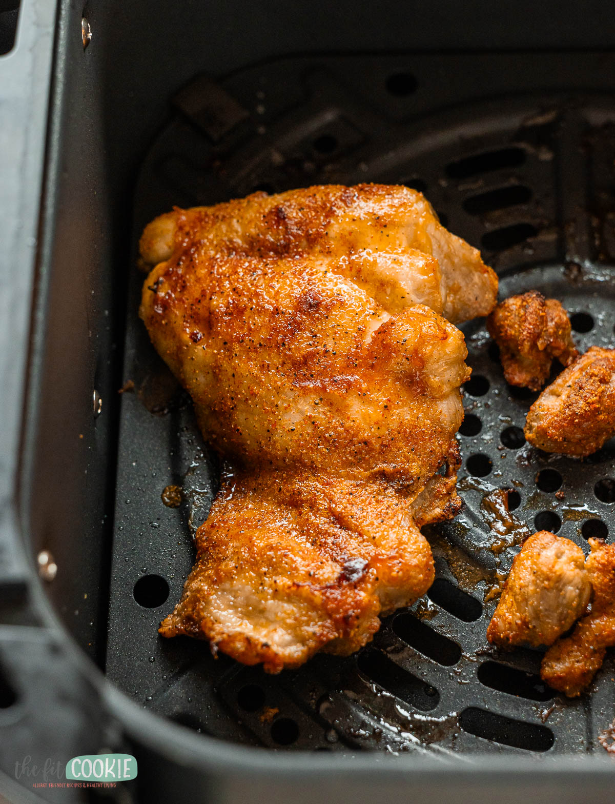 A cooked, seasoned air fryer chicken thigh sits in the basket with a few small pieces of chicken nearby.