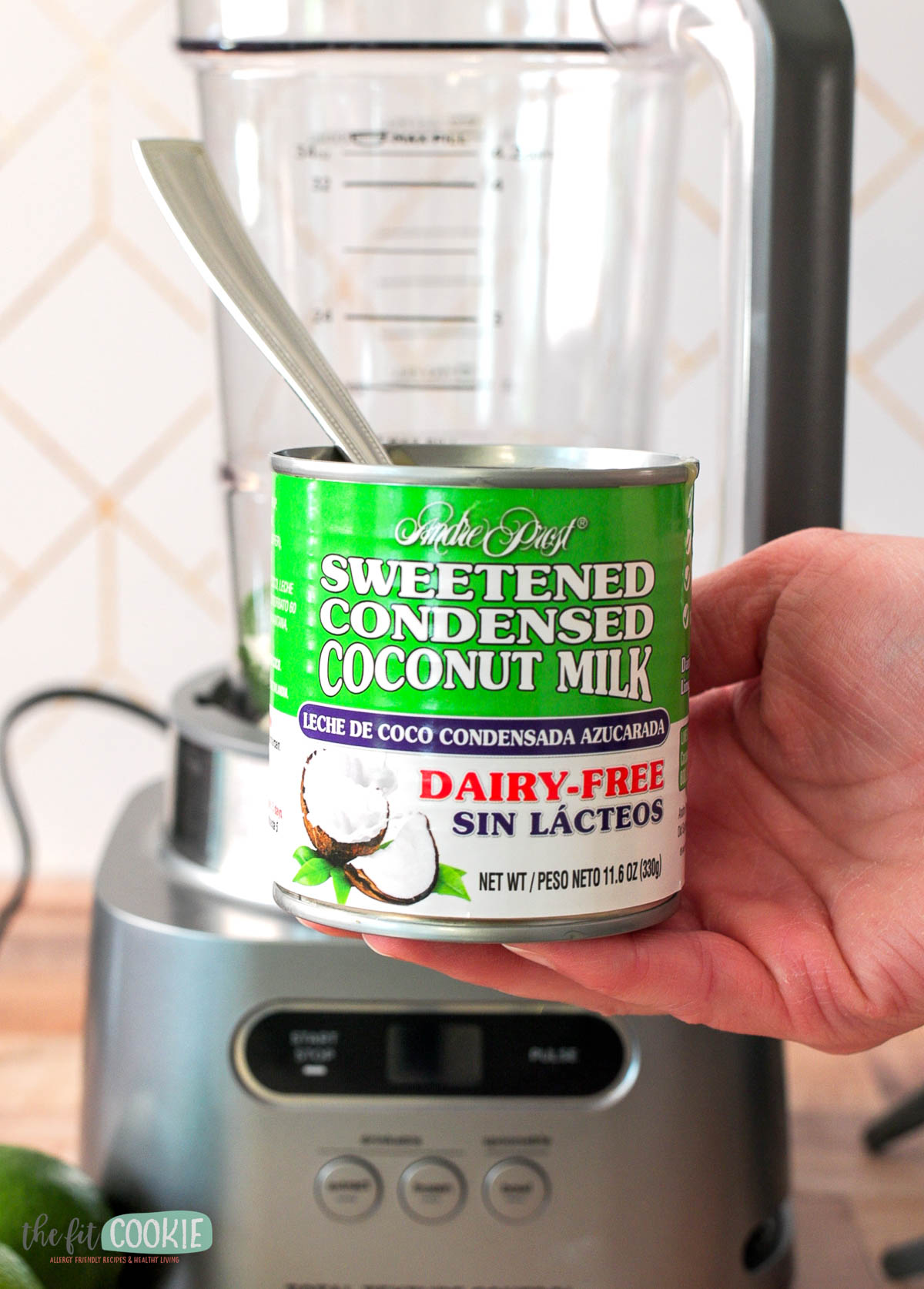 A hand holds a can of dairy-free sweetened condensed coconut milk in front of a blender.