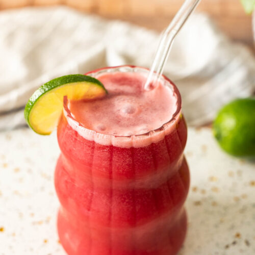 A glass of dairy free red watermelon drink with a glass straw and a lime slice garnish sits on a white countertop, offering a refreshing twist reminiscent of Brazilian Lemonade.