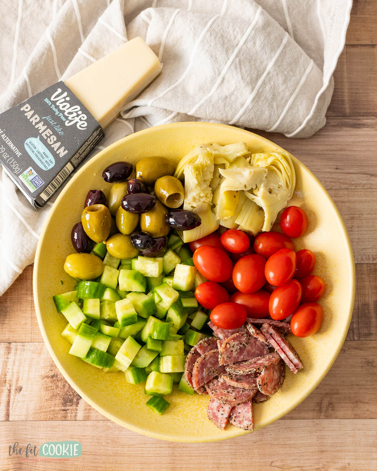 A yellow plate with chopped cucumber, cherry tomatoes, artichoke hearts, salami slices, and marinated olives.