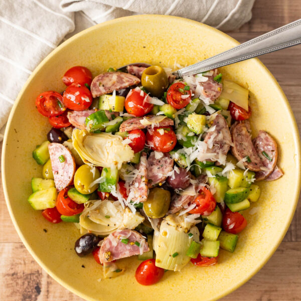 A yellow bowl of Italian Chopped Salad with sausage, cherry tomatoes, cucumbers, olives, artichoke hearts, and shredded cheese sits on a wooden table alongside a fork and a striped napkin—an easy salad perfect for any occasion.