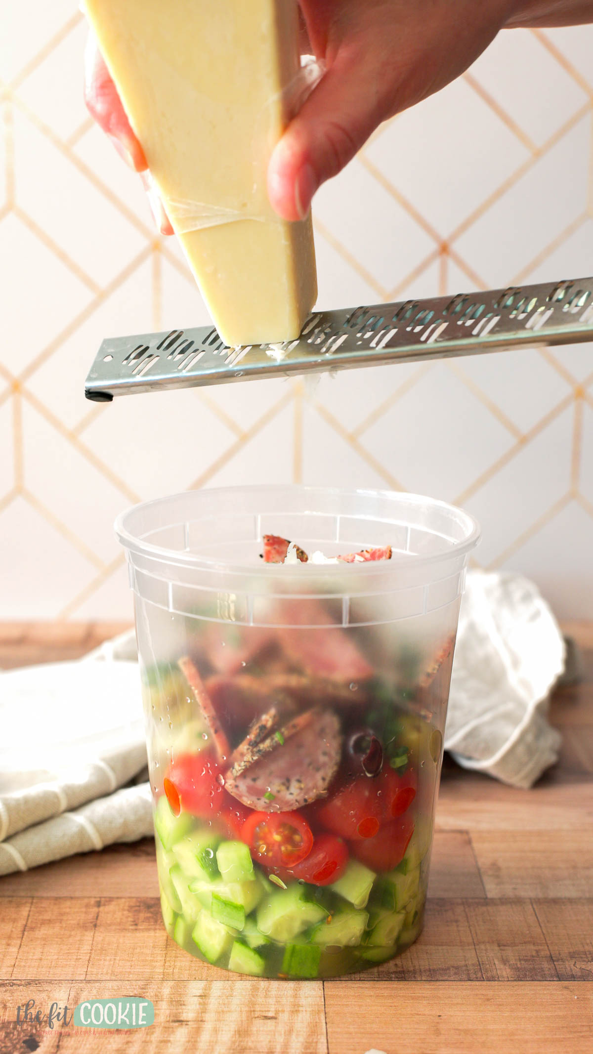 A hand grates dairy free parmesan cheese over a plastic container filled with Italian chopped salad.