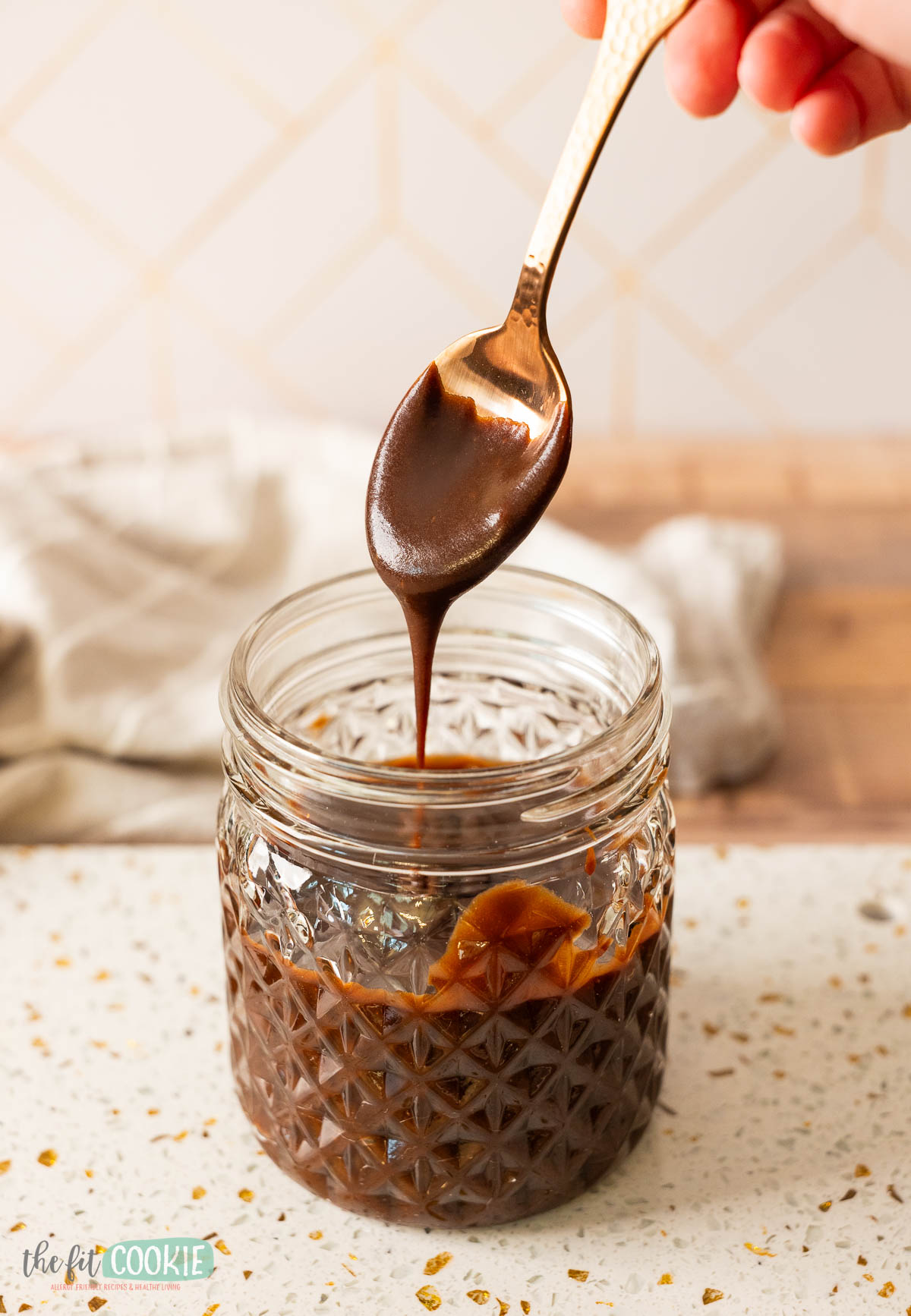 A hand holds a spoon over a glass jar filled with thick, vegan hot fudge sauce.