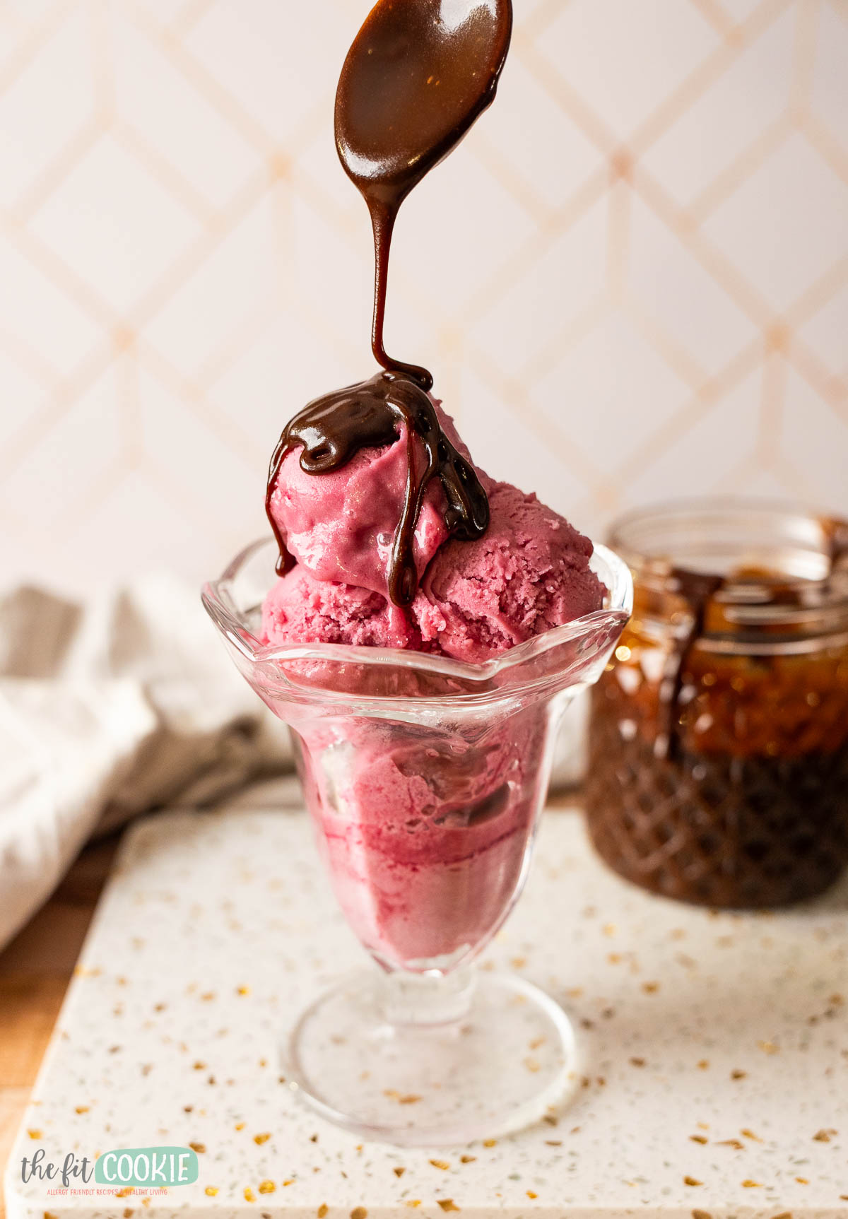 A spoon drizzles Dairy Free Hot Fudge over scoops of berry ice cream in a glass dish.