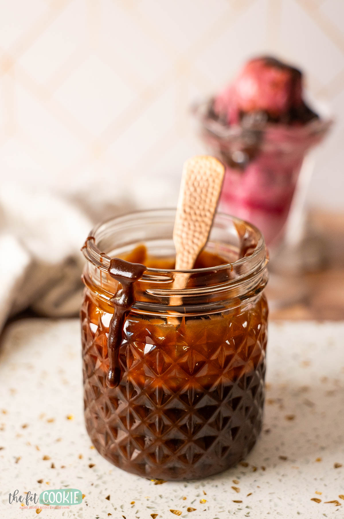 A glass jar filled with dairy free hot fudge sauce sits on a countertop, with a spoon inside and some sauce dripping down the side.