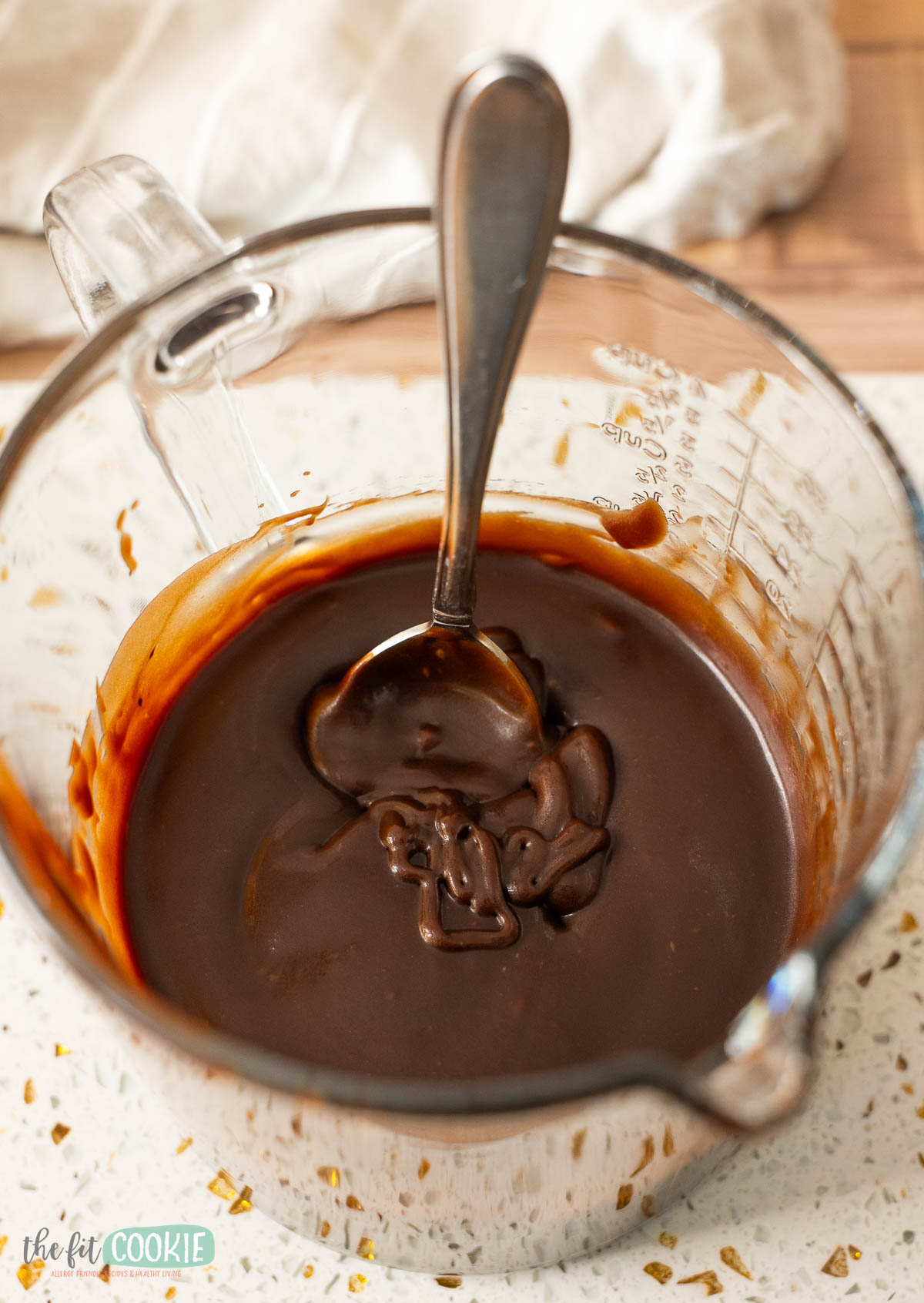 A glass measuring cup filled with thick, Dairy free hot fudge sauce with a spoon inside.