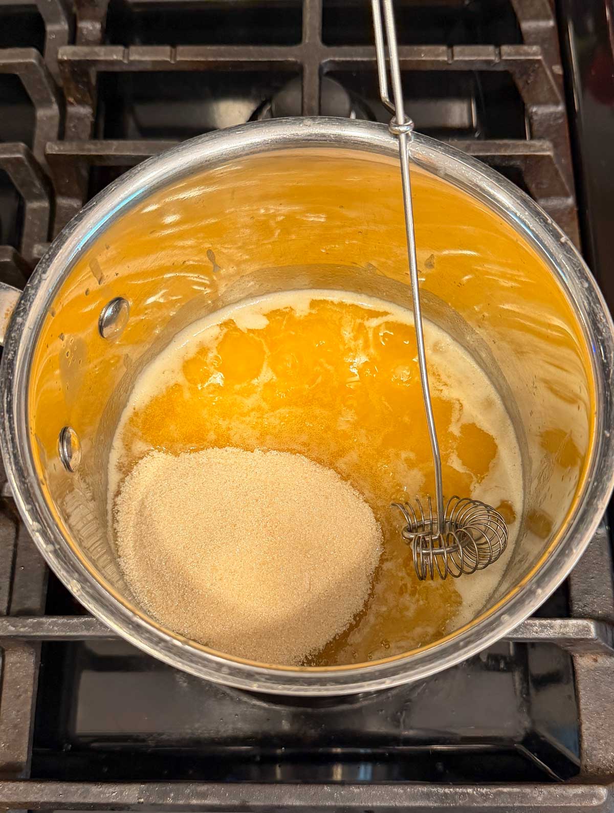 A saucepan with melted butter and sugar sits on a stove top, with a metal whisk resting inside the pot.